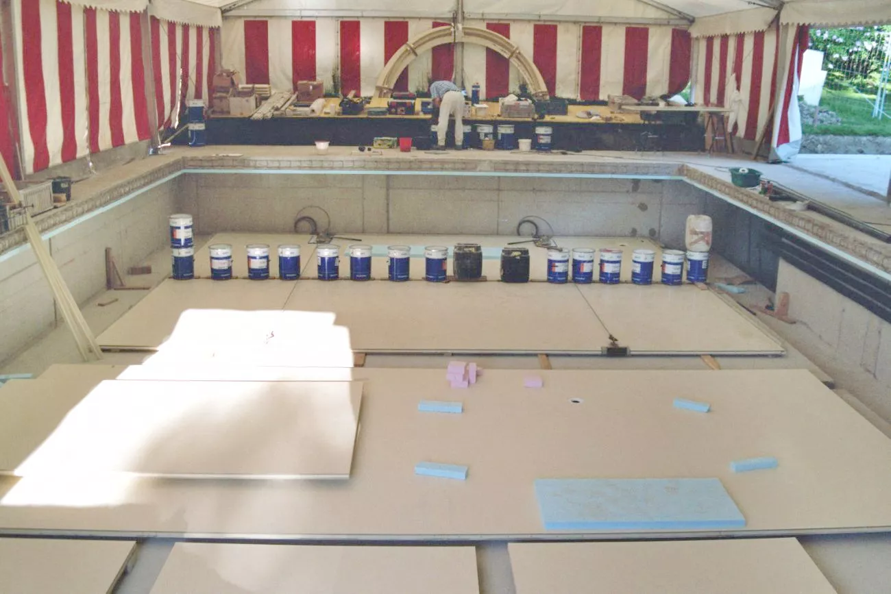 Das Bild zeigt einen Arbeiter bei einem Schwimmbecken, das mit einem Festzelt gegen Regen geschützt ist. Auf dem Boden das Schwimmbeckens sind schon Beckensegmente am Boden angebracht und es stehen GFK-Fässer zur Verarbeitung bereit.